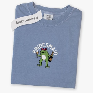 May include: A blue crew neck t-shirt with an embroidered graphic of a green frog wearing a pink headband, holding a champagne glass and a bottle. The text "BRIDESMAID" is arched above the frog in white. The shirt has a Comfort Colors label.
