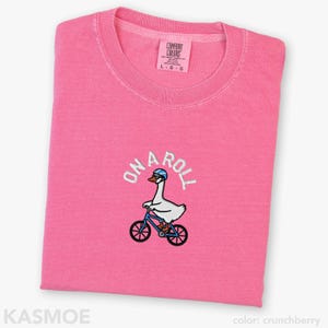 May include: A bright pink short-sleeved t-shirt featuring an embroidered graphic of a white goose wearing a blue helmet and sunglasses while riding a blue bicycle. The text ON A ROLL is embroidered in white above the goose. The shirt has a crew neckline.
