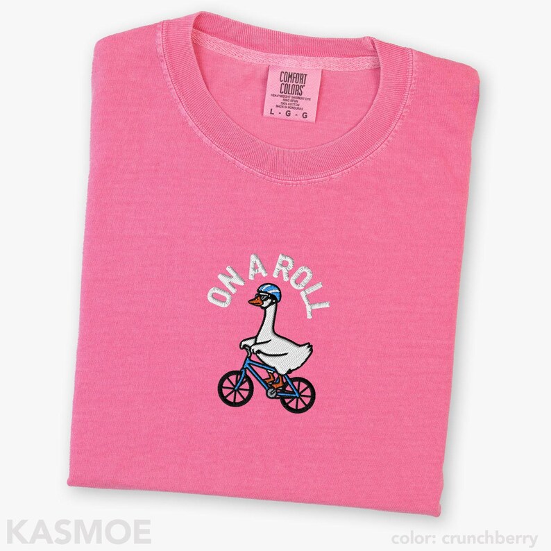 May include: A bright pink short-sleeved t-shirt featuring an embroidered graphic of a white goose wearing a blue helmet and sunglasses while riding a blue bicycle. The text ON A ROLL is embroidered in white above the goose. The shirt has a crew neckline.