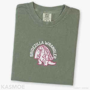 Może przedstawiać: T-shirt z okrągłym dekoltem w kolorze mchu z haftem przedstawiającym stworzenie w welonie ślubnym. Napis BRIDEZILLA WRANGLER jest wyhaftowany białymi nićmi w łuk nad stworzeniem. Metka Comfort Colors wskazuje rozmiar L.