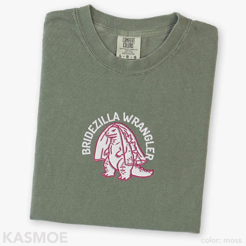 Może przedstawiać: T-shirt z okrągłym dekoltem w kolorze mchu z haftem przedstawiającym stworzenie w welonie ślubnym. Napis BRIDEZILLA WRANGLER jest wyhaftowany białymi nićmi w łuk nad stworzeniem. Metka Comfort Colors wskazuje rozmiar L.