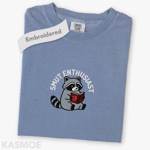 May include: A blue crewneck t-shirt featuring an embroidered graphic of a raccoon holding a red book. The text "SMUT ENTHUSIAST" is arched above the raccoon in white thread. The shirt has a Comfort Colors label at the inner neckline.