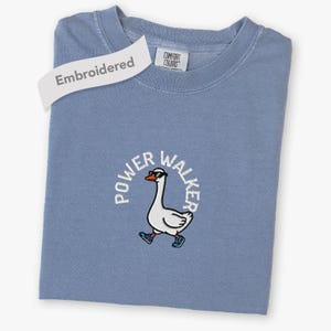 May include: A blue crew neck t-shirt featuring an embroidered graphic of a white goose wearing black sunglasses and blue sneakers. The text "POWER WALKER" is arched above the goose in white lettering. A tag reads "COMFORT COLORS".