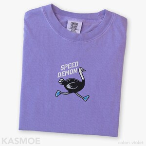 May include: A violet short-sleeve t-shirt featuring an embroidered graphic of a black ostrich running while wearing blue sneakers. Above the ostrich, the text SPEED DEMON is embroidered in white. The inner neck label reads COMFORT COLORS.