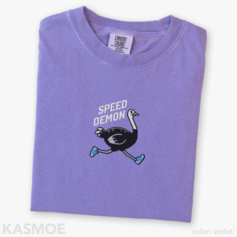 May include: A violet short-sleeve t-shirt featuring an embroidered graphic of a black ostrich running while wearing blue sneakers. Above the ostrich, the text SPEED DEMON is embroidered in white. The inner neck label reads COMFORT COLORS.