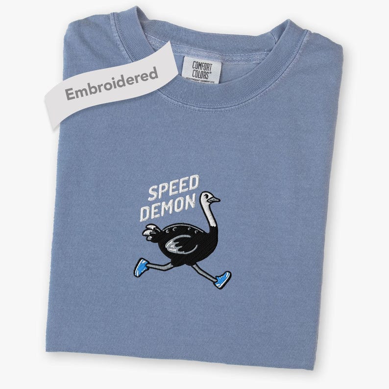 May include: A blue short-sleeved t-shirt featuring an embroidered graphic of a black ostrich running while wearing blue sneakers. The text "SPEED DEMON" is embroidered in white above the ostrich. The shirt has a Comfort Colors brand label.