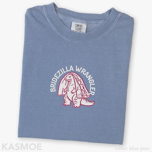 Może przedstawiać: Dżinsowoniebieski t-shirt z haftowaną grafiką przedstawiającą stworzenie w welonie ślubnym. Napis BRIDEZILLA WRANGLER jest wyhaftowany białymi nićmi w łuk nad stworzeniem. Koszulka posiada metkę marki Comfort Colors przy kołnierzyku.