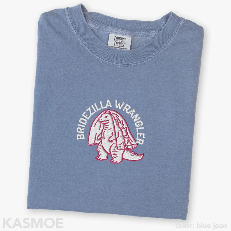 Może przedstawiać: Dżinsowoniebieski t-shirt z haftowaną grafiką przedstawiającą stworzenie w welonie ślubnym. Napis BRIDEZILLA WRANGLER jest wyhaftowany białymi nićmi w łuk nad stworzeniem. Koszulka posiada metkę marki Comfort Colors przy kołnierzyku.