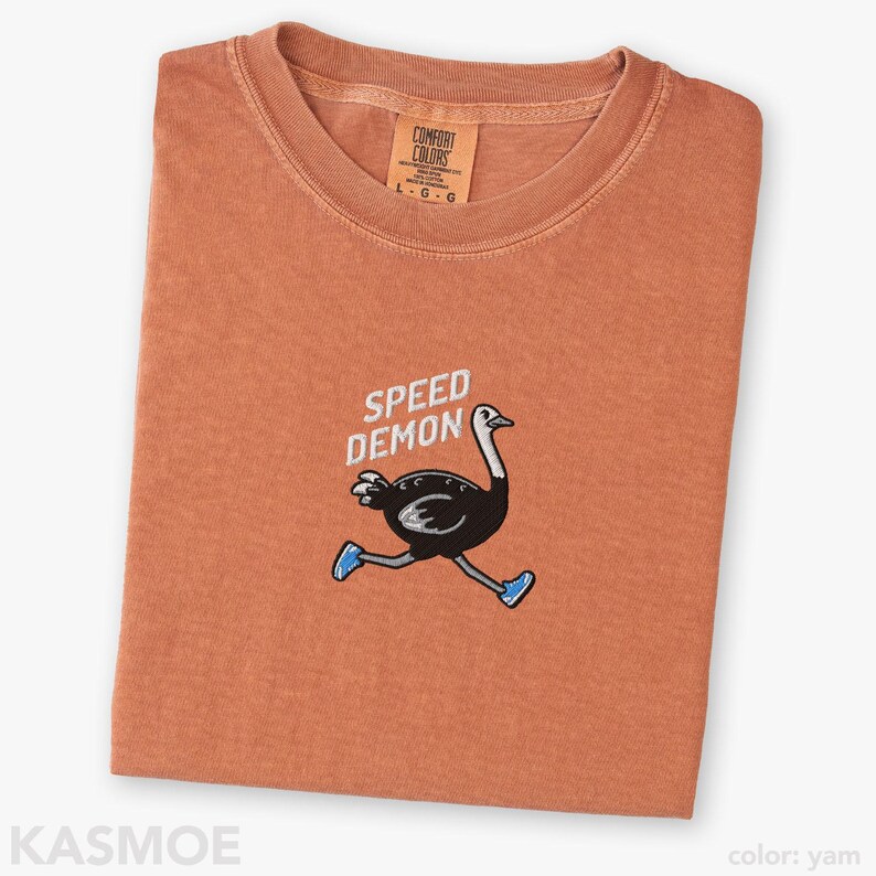 May include: A yam-colored t-shirt featuring an embroidered graphic of a black ostrich running while wearing blue sneakers. Above the ostrich, the text SPEED DEMON is embroidered in white. The shirt has a crew neckline and a Comfort Colors brand label.
