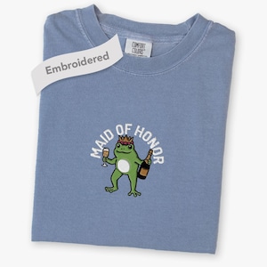 Könnte beinhalten: Ein blaues T-Shirt mit einer Stickerei eines grünen Frosches, der eine Krone trägt und ein Champagnerglas sowie eine Flasche hält. Der Text "MAID OF HONOR" ist darüber gebogen. Ein weißes Etikett zeigt "Embroidered" und das Nackenlabel "COMFORT COLORS".