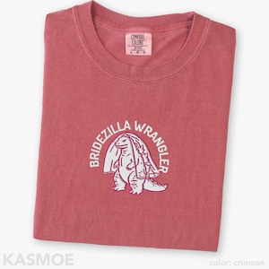 Może przedstawiać: Karmazynowy t-shirt z okrągłym dekoltem i metką marki Comfort Colors na wewnętrznej stronie kołnierza. Na piersi znajduje się biały haft przedstawiający stworzenie w welonie, otoczone łukowatym napisem BRIDEZILLA WRANGLER pogrubioną czcionką.