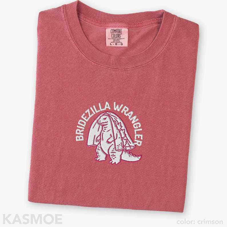 Może przedstawiać: Karmazynowy t-shirt z okrągłym dekoltem i metką marki Comfort Colors na wewnętrznej stronie kołnierza. Na piersi znajduje się biały haft przedstawiający stworzenie w welonie, otoczone łukowatym napisem BRIDEZILLA WRANGLER pogrubioną czcionką.