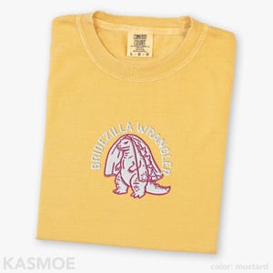 Może przedstawiać: Musztardowy t-shirt z okrągłym dekoltem i haftowaną grafiką przedstawiającą dinozaura w welonie ślubnym. Napis BRIDEZILLA WRANGLER jest wyhaftowany białymi nićmi w łuk nad stworzeniem. Koszulka posiada metkę marki Comfort Colors przy dekolcie.