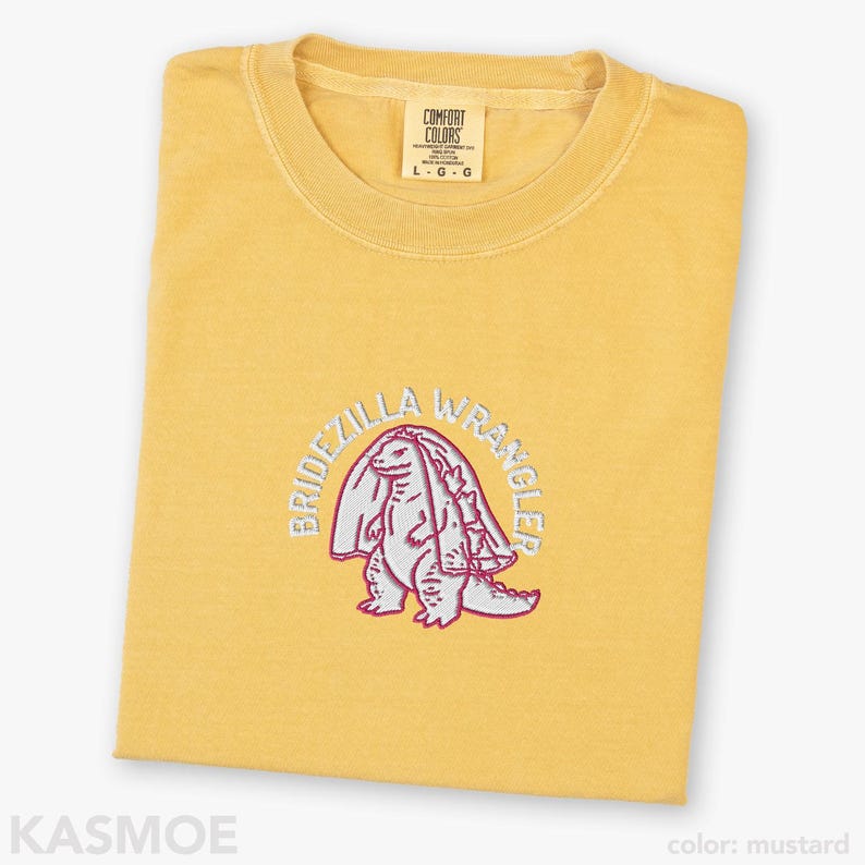 Może przedstawiać: Musztardowy t-shirt z okrągłym dekoltem i haftowaną grafiką przedstawiającą dinozaura w welonie ślubnym. Napis BRIDEZILLA WRANGLER jest wyhaftowany białymi nićmi w łuk nad stworzeniem. Koszulka posiada metkę marki Comfort Colors przy dekolcie.