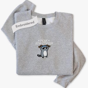 May include: A light gray crewneck sweatshirt featuring an embroidered graphic of a raccoon wearing a blue headband and lifting two brown dumbbells. Above the raccoon, the text reads PEAK ATHLETICISM with a star on each side of the word PEAK.