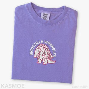 Może przedstawiać: Fioletowy t-shirt z kr&oacute;tkim rękawem z wyhaftowaną grafiką przedstawiającą dinozaura w welonie ślubnym. Napis "BRIDEZILLA WRANGLER" jest wygięty w łuk nad dinozaurem białą nicią. Koszulka ma okrągły dekolt i metkę marki Comfort Colors.