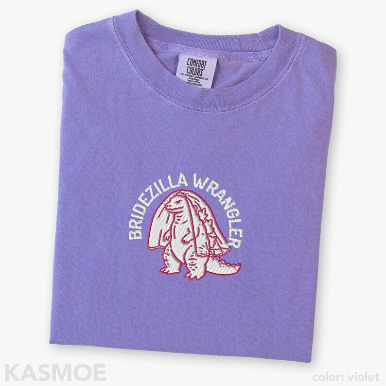 Może przedstawiać: Fioletowy t-shirt z kr&oacute;tkim rękawem z wyhaftowaną grafiką przedstawiającą dinozaura w welonie ślubnym. Napis "BRIDEZILLA WRANGLER" jest wygięty w łuk nad dinozaurem białą nicią. Koszulka ma okrągły dekolt i metkę marki Comfort Colors.