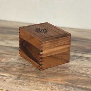 May include: A wooden box with a hinged lid, constructed with dovetail joints. The box is made of a warm-toned wood and features an engraved floral design on the lid. The box is sitting on a wooden surface.