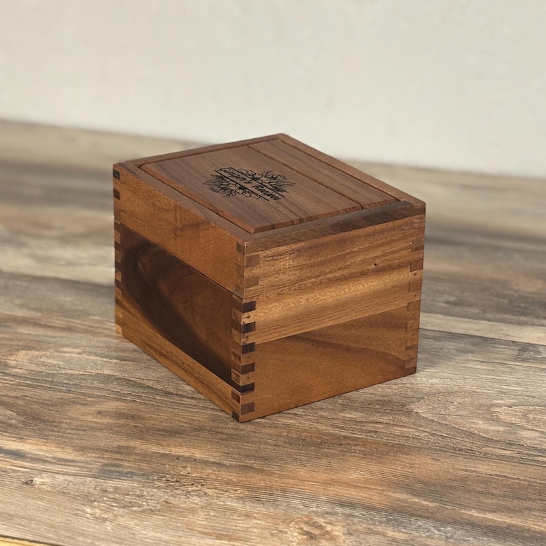May include: A wooden box with a hinged lid, constructed with dovetail joints. The box is made of a warm-toned wood and features an engraved floral design on the lid. The box is sitting on a wooden surface.