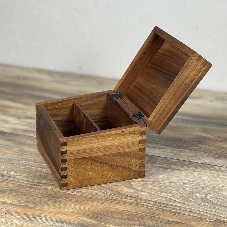May include: A wooden box with a hinged lid, open to reveal two interior compartments. The box is constructed with dovetail joints and has a natural wood grain finish. The lid is partially open, showing the interior.