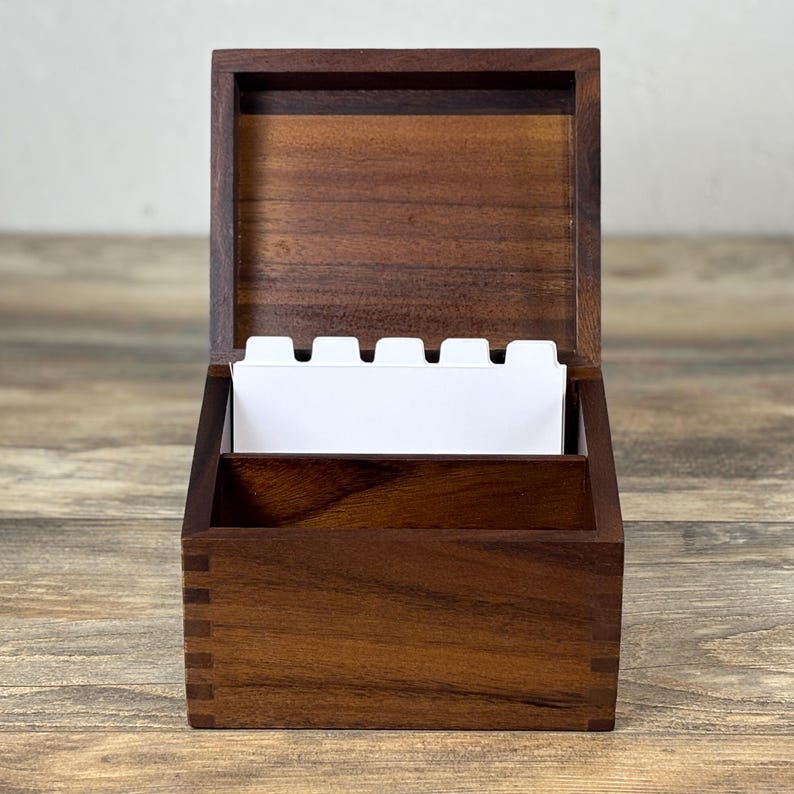 May include: A wooden card box with a hinged lid, open to reveal white index cards. The box is made of dark brown wood with visible grain and dovetail joints. The box is sitting on a wooden surface.