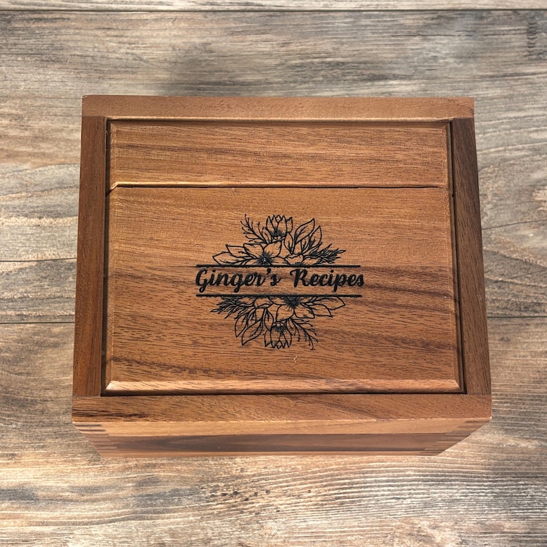 May include: A wooden recipe box with a hinged lid. The box is engraved with the words "Ginger's Recipes" surrounded by a floral design. The box is made of a warm-toned wood and has a rectangular shape.