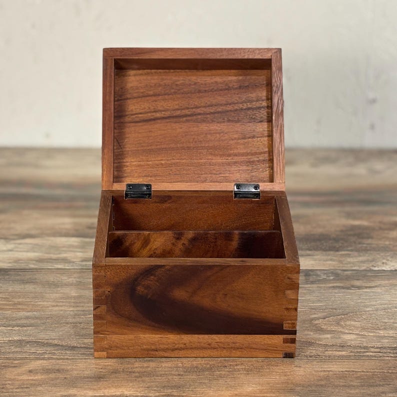 May include: Open wooden box with a hinged lid. The box is made of dark brown wood with visible grain and is divided into two compartments. The hinges are black. The box is sitting on a wooden surface.