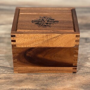 May include: A wooden recipe box with a hinged lid. The box is a warm brown color and features dovetail joints. The lid is engraved with the words "Ginger's Recipes" in a decorative font, with a floral design. The box is sitting on a wooden surface.