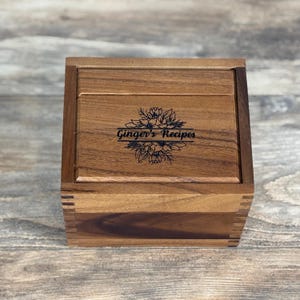 May include: A wooden recipe box with a hinged lid. The box is made of a light brown wood and features a floral design with the text "Ginger's Recipes" engraved on the lid. The box is square-shaped and has a dovetail joint construction.