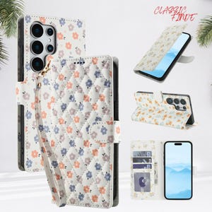 Pode incluir: Capa de telefone floral branca com design acolchoado. A capa possui uma alça de pulso e um fecho magnético. O design inclui pequenas flores em tons de laranja, azul e roxo. O texto "CLASSIC FINDE" é visível.