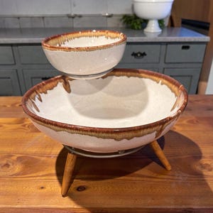 May include: Two ceramic serving bowls. The top bowl is smaller and sits above a larger bowl. Both bowls have a white interior and a brown, dripped glaze around the rim. The larger bowl rests on a metal stand with wooden legs.