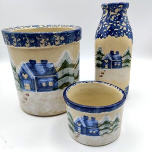 May include: A set of three ceramic pieces: a large planter, a small bowl, and a tall vase. Each piece is painted with a winter scene featuring blue houses, snow, and trees against a cream background, with blue rims.
