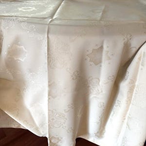 Puede incluir: Mantel de color marfil con un sutil patrón floral y de estrella repetido. La tela tiene un ligero brillo y el diseño está en relieve, creando una superficie texturizada. La tela está drapeada, mostrando pliegues y sombras.
