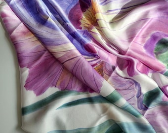 May include: A silk scarf with a floral iris design. The scarf features large, detailed iris blooms in shades of purple, pink, and yellow against a white background. Green and blue stripes accent the edges. The fabric has a soft sheen.