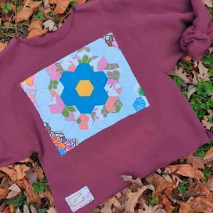 May include: A maroon long-sleeve sweatshirt featuring a large, square, quilted patch on the front. The patch displays a central blue hexagonal flower with a yellow center, surrounded by light blue, pink, and green floral-patterned fabric hexagons.