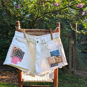Puede incluir: Shorts vaqueros blancos con bordes deshilachados y detalles de patchwork coloridos, colgados en una silla de madera al aire libre. Los parches incluyen telas rosas, azules y a cuadros en blanco y negro. Los shorts se sujetan con pinzas de madera.