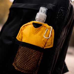May include: A yellow water bottle in a black mesh pocket, attached to a black backpack. The bottle has a clear spray nozzle and a silver carabiner clip. The bottle is shaped like a yellow shirt.