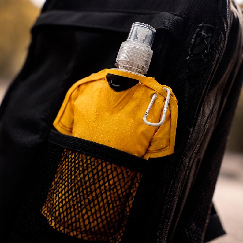 May include: A yellow water bottle in a black mesh pocket, attached to a black backpack. The bottle has a clear spray nozzle and a silver carabiner clip. The bottle is shaped like a yellow shirt.