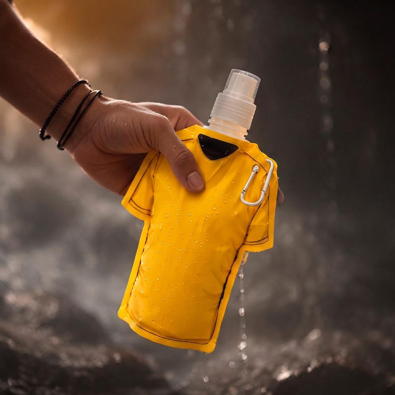 May include: A yellow water bottle shaped like a jersey, held by a hand. The bottle has a white spray nozzle and a small black accent. A silver carabiner clip is attached. Water droplets are visible on the bottle.