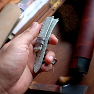 May include: A light gray leather watch strap with a textured surface. The strap has a silver-colored buckle and is held in a person's hand. The background is a wooden surface with tools and a leather container.