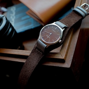 Op de afbeelding: Een bruin polshorloge met een zilveren kast en een bruine leren band. De wijzerplaat van het horloge is bruin met zilveren wijzers en markeringen. Het horloge ligt op een houten oppervlak.