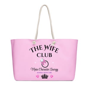 May include: A large pink tote bag with white rope handles. The bag features the text "THE WIFE CLUB" in black script, along with "Main Character Energy" and a ring graphic. Two pink hearts and a crown are also visible.