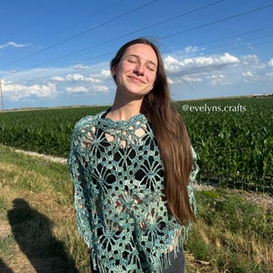 May include: A teal and green crocheted poncho with a floral openwork pattern and fringing. The garment is shown outdoors, set against a blue sky and a green field.