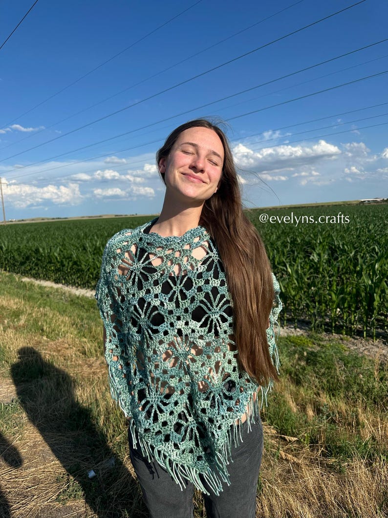 May include: A teal and green crocheted poncho with a floral openwork pattern and fringing. The garment is shown outdoors, set against a blue sky and a green field.