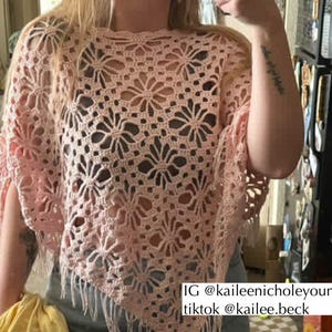 May include: A light pink crochet poncho with a floral design. The poncho has a fringed hemline and is made of a soft, open-weave material. The image includes the text "IG @kaileenicholeyoung tiktok @kailee.beck".