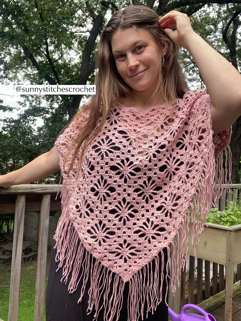 May include: A light pink crochet poncho with a floral pattern and fringe. The poncho is handmade and features a V-neck design. The text "@sunnystitchescrochet" is visible in the image. This item is perfect for adding a touch of bohemian style to any outfit.