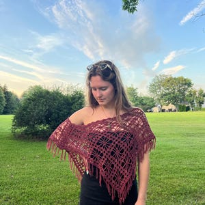 May include: A burgundy crochet shawl with a floral pattern and fringe. The shawl is worn over a black top. The background features green grass and a blue sky.