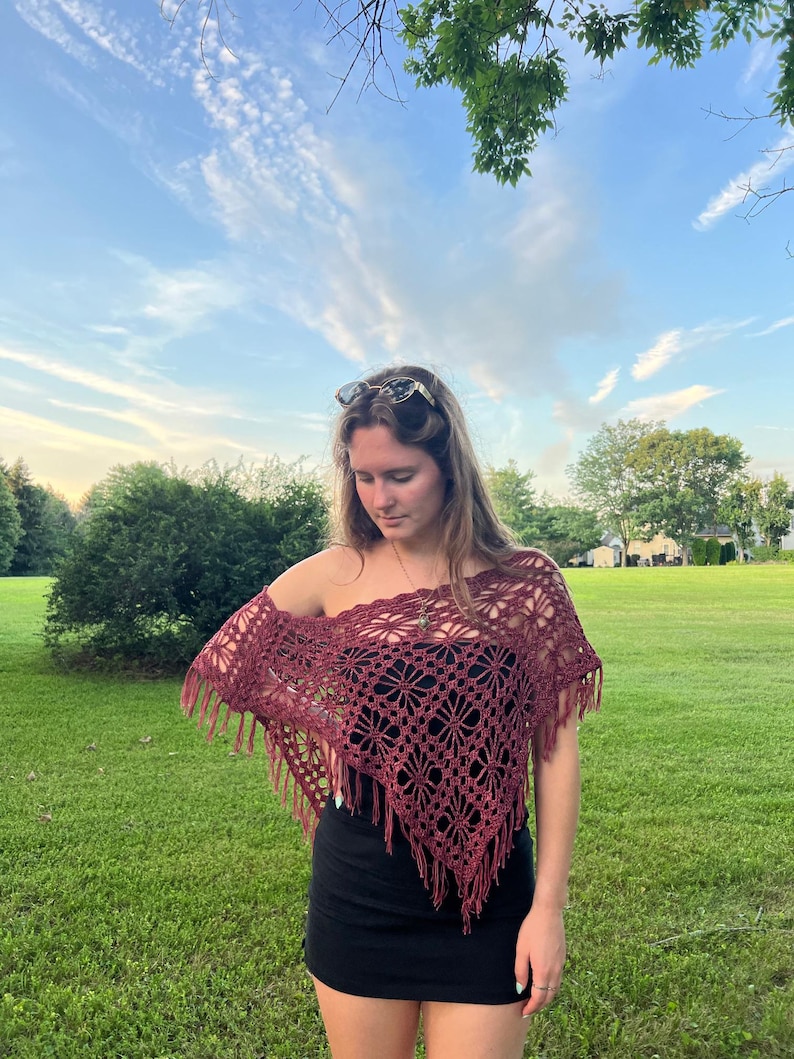 May include: A burgundy crochet shawl with a floral pattern and fringe. The shawl is worn over a black top. The background features green grass and a blue sky.