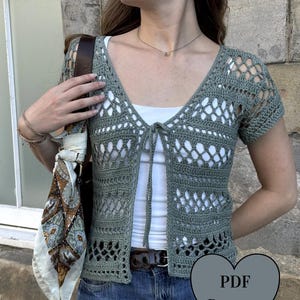 May include: A sage green short-sleeved crochet cardigan featuring an open-front design with thin tie closures, a V-neckline, and a repeating diamond-shaped lattice pattern. A heart-shaped graphic in the corner displays the text PDF Pattern.