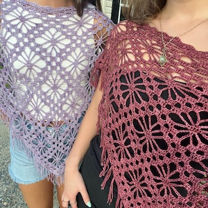 May include: Two crochet ponchos are shown. One is a light lilac colour, and the other is a deep red-purple. Both have a floral pattern and fringe along the bottom. The ponchos are worn over other clothing.